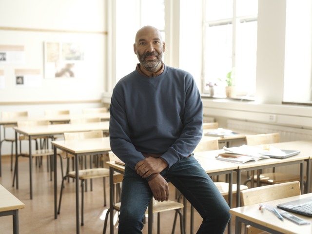 Manlig gymnasielärare halvsitter på en bänk i ett klassrum Gymnasieskola - porträtt