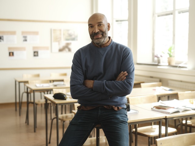 Leende manlig gymnasielärare halvsitter på en bänk i ett klassrum Gymnasieskola - porträtt