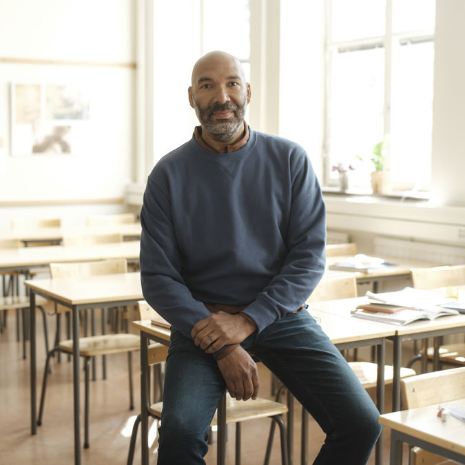 Manlig gymnasielärare halvsitter på en bänk i ett klassrum.