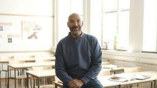 Manlig gymnasielärare halvsitter på en bänk i ett klassrum.