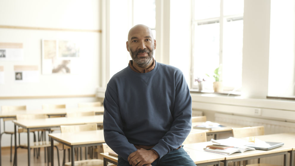Manlig gymnasielärare halvsitter på en bänk i ett klassrum.