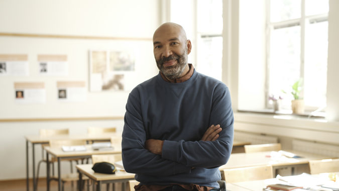 Leende manlig gymnasielärare halvsitter på en bänk i ett klassrum.