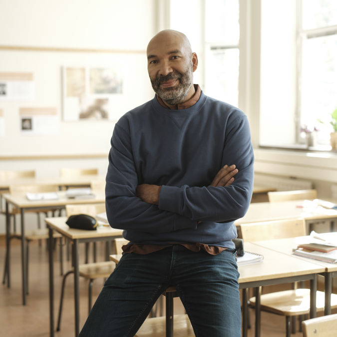 Leende manlig gymnasielärare halvsitter på en bänk i ett klassrum.