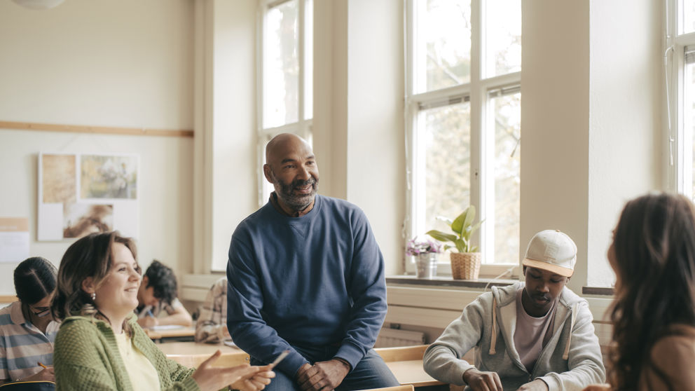 Leende gymnasielärare i samtal med elever under lektion.