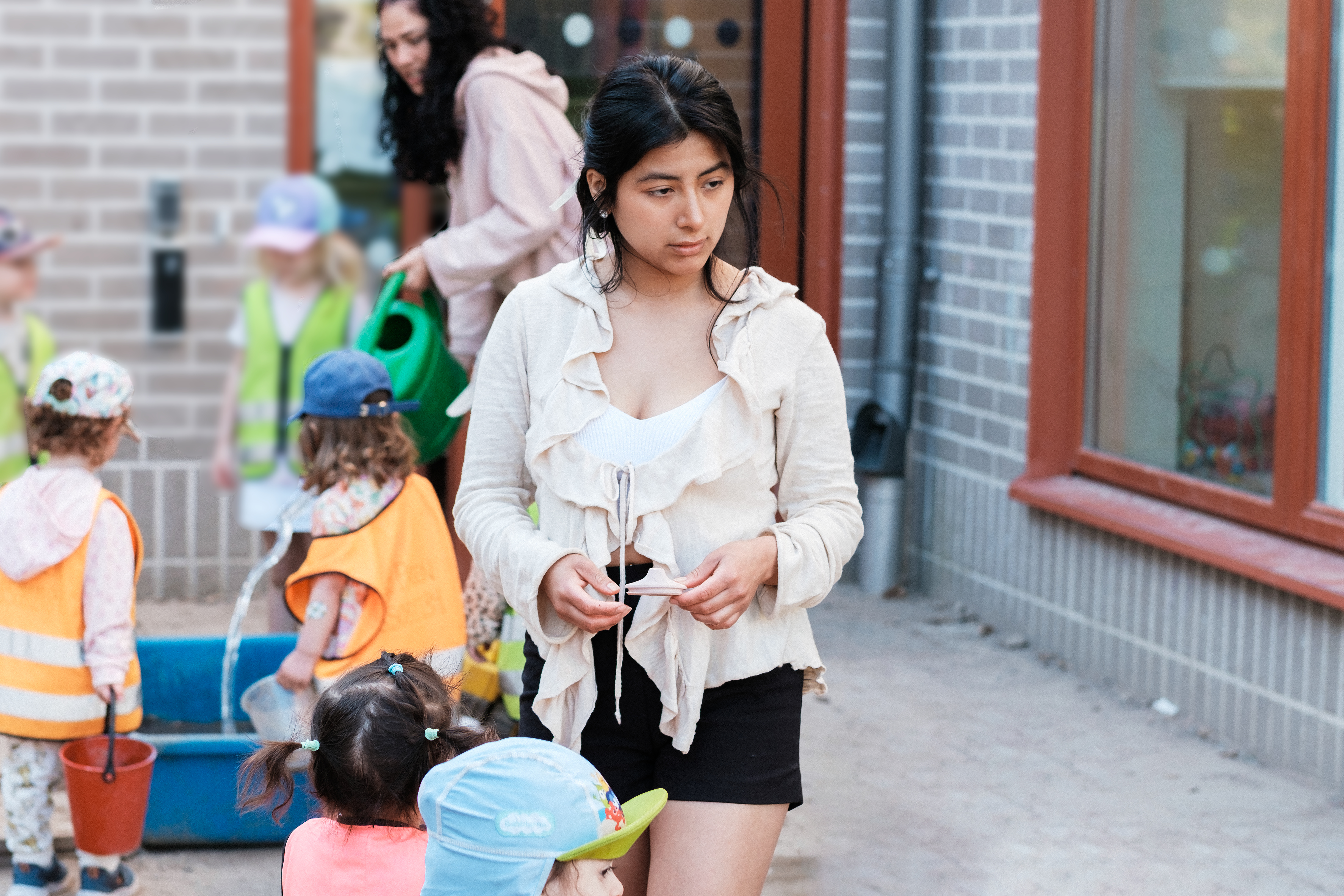 Ung, kvinnlig förskollärare står utomhus och verkar vara i sina egna tankar, med barn i bakgrunden.