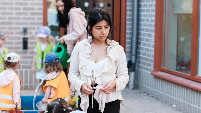 Ung, kvinnlig förskollärare står utomhus och verkar vara i sina egna tankar, med barn i bakgrunden.