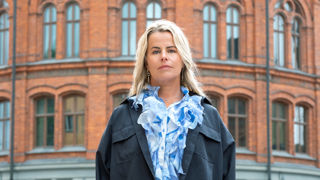 Anna Olskog med neutral min, stående utomhus iklädd mörkblå jacka och en ljusblå blus med en massa krås/volanger.