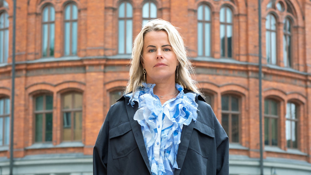 Anna Olskog med neutral min, stående utomhus iklädd mörkblå jacka och en ljusblå blus med en massa krås/volanger.