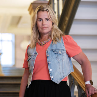 Anna Olskog med neutral min, stående i trappa inomhus. Hon är iklädd en aprikosfärgad t-shirt och kort jeansjacka.