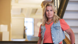 Anna Olskog med neutral min, stående i trappa inomhus. Hon är iklädd en aprikosfärgad t-shirt och kort jeansjacka.