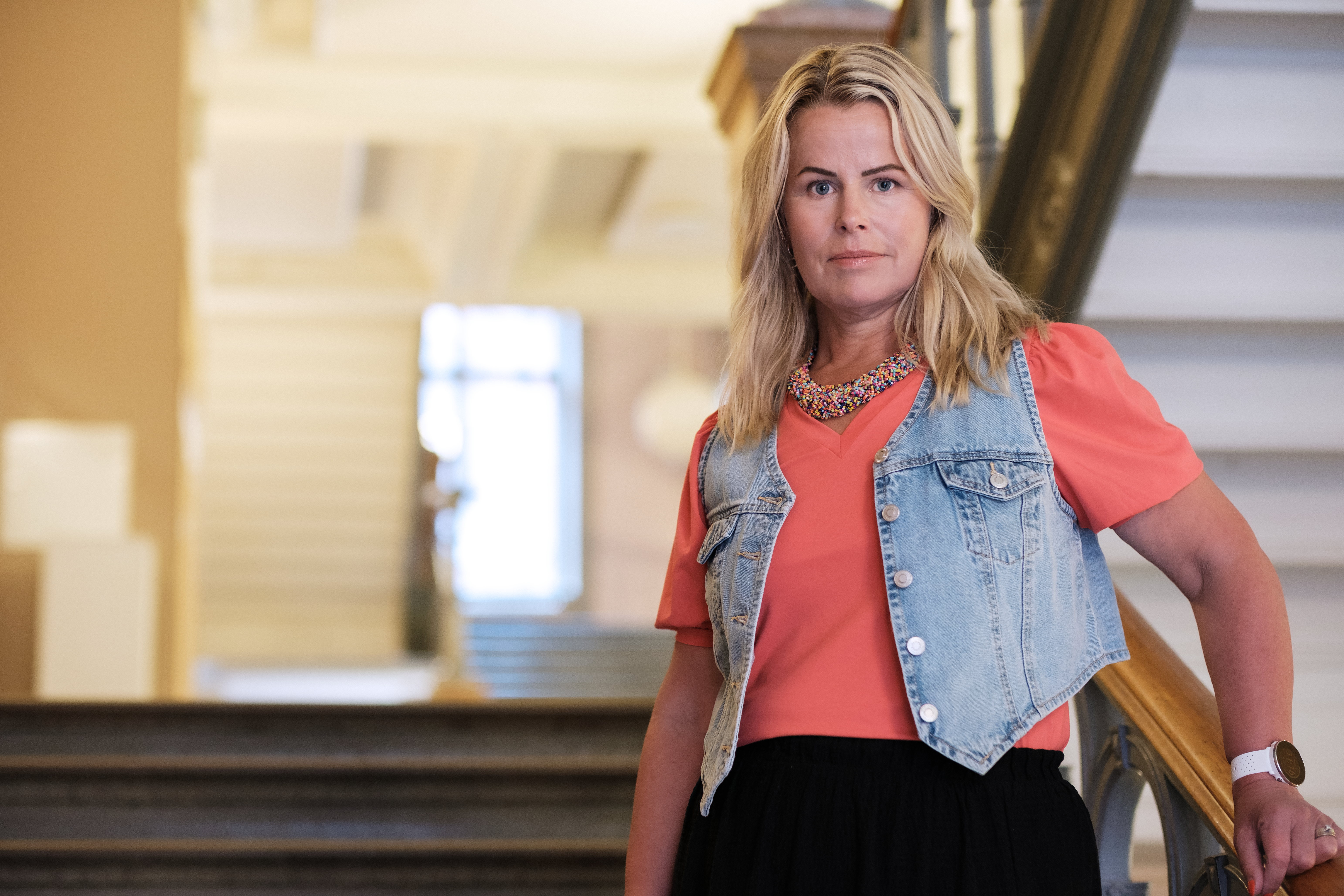 Anna Olskog med neutral min, stående i trappa inomhus. Hon är iklädd en aprikosfärgad t-shirt och kort jeansjacka.