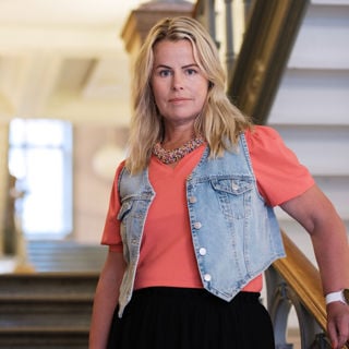 Anna Olskog med neutral min, stående i trappa inomhus. Hon är iklädd en aprikosfärgad t-shirt och kort jeansjacka.