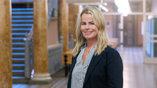 Leende Anna Olskog i en tom skolkorridor, iklädd mörk kavaj och en blårandig blus.