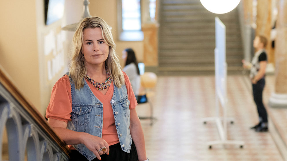 Anna Olskog med neutral min iklädd aprikosfärgad t-shirt och kort jeansjacka, stående i en trappa inomhus med ett par personer i bakgrunden.