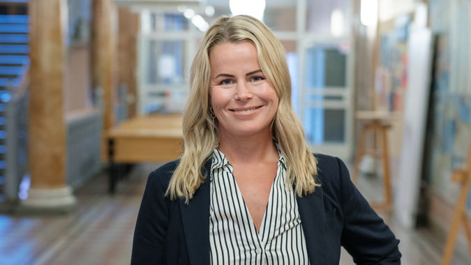 Leende Anna Olskog stående i en tom skolkorridor, där hon har mörk kavaj och en blårandig blus.