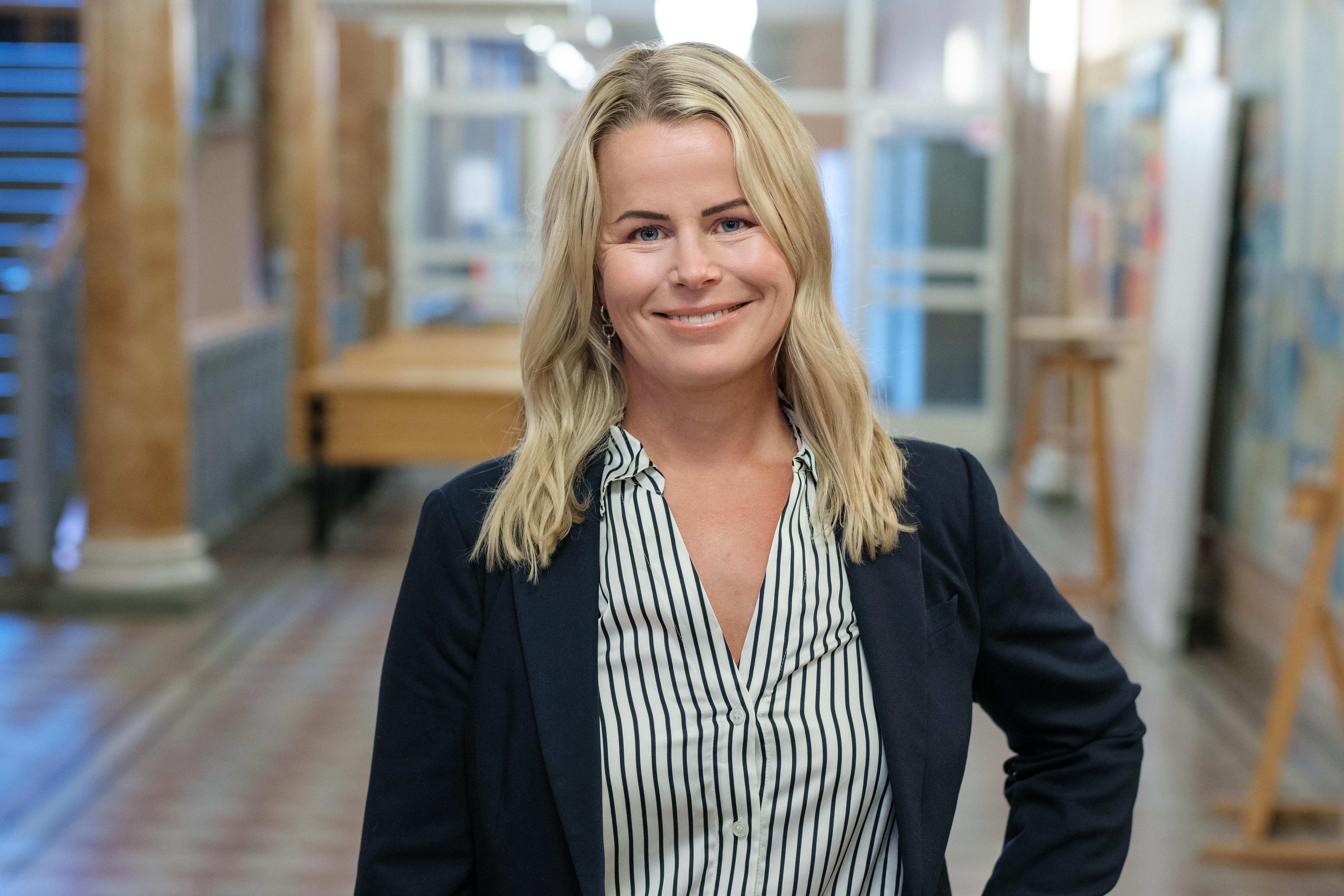 Leende Anna Olskog stående i en tom skolkorridor, där hon har mörk kavaj och en blårandig blus.