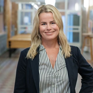 Leende Anna Olskog stående i en tom skolkorridor, där hon har mörk kavaj och en blårandig blus.