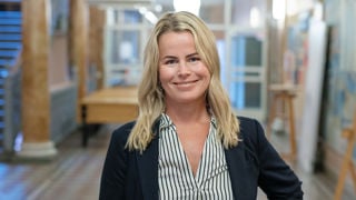 Leende Anna Olskog stående i en tom skolkorridor, där hon har mörk kavaj och en blårandig blus.