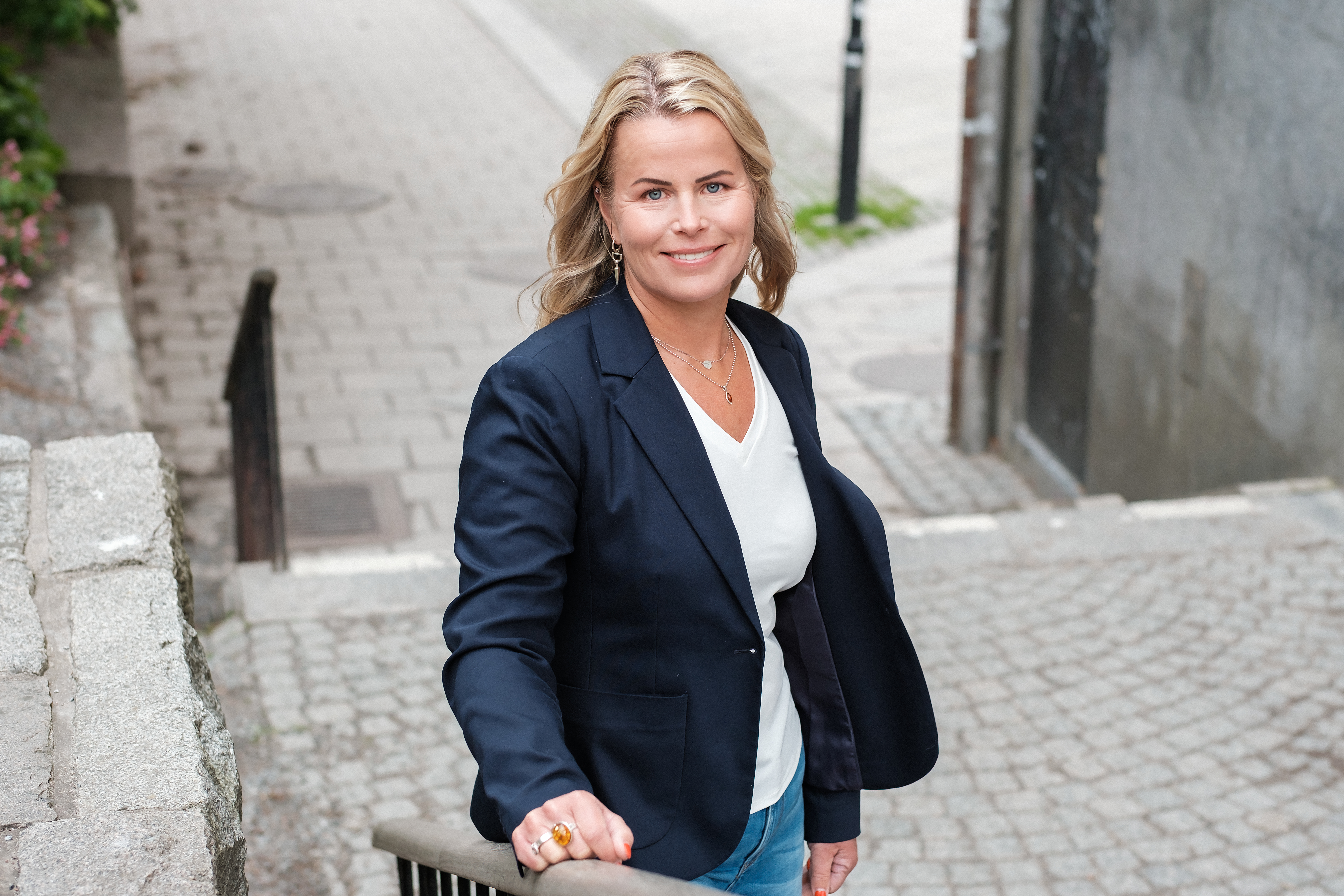 Leende Anna Olskog, iklädd vit t-shirt och mörk kavaj, stående i trappa utomhus.