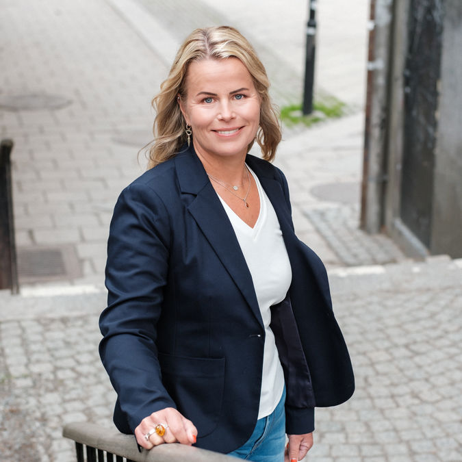 Leende Anna Olskog, iklädd vit t-shirt och mörk kavaj, stående i trappa utomhus.
