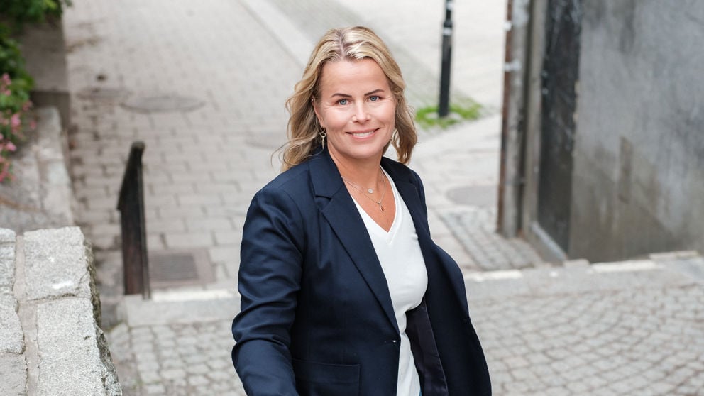 Leende Anna Olskog, iklädd vit t-shirt och mörk kavaj, stående i trappa utomhus.
