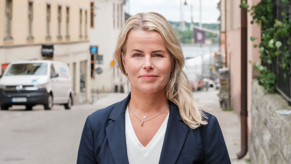Lätt leende Anna Olskog stående på en tom gata utomhus, där hon har på sig vit t-shirt och mörkblå kavaj.