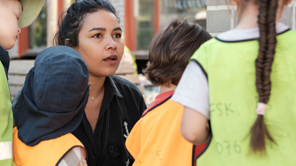 Kvinnlig, ung förskollärare pratar med barn i reflexvästar utomhus på Påsens förskola.