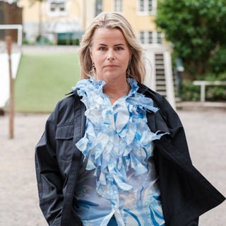 Sveriges Lärares ordförande Anna Olskog med en neutral min står utomhus på sommarhalvåret, klädd i ljusblå blus med en massa krås/volanger och en mörk jacka.