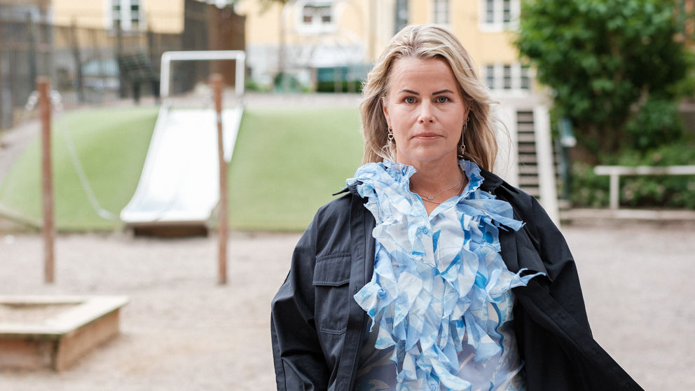 Sveriges Lärares ordförande Anna Olskog med en neutral min står utomhus på sommarhalvåret, klädd i ljusblå blus med en massa krås/volanger och en mörk jacka.