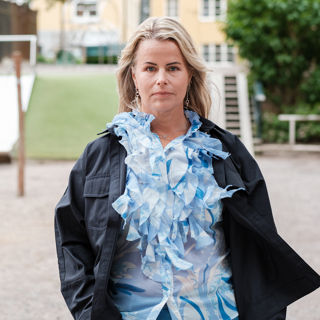 Sveriges Lärares ordförande Anna Olskog med en neutral min står utomhus på sommarhalvåret, klädd i ljusblå blus med en massa krås/volanger och en mörk jacka.