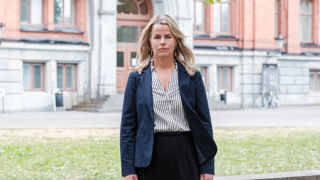 Sveriges Lärares ordförande Anna Olskog står utomhus på sommarhalvåret. Hon har neutral, nästan sur min och är klädd i vit blus med mörka ränder och en mörk kavaj.