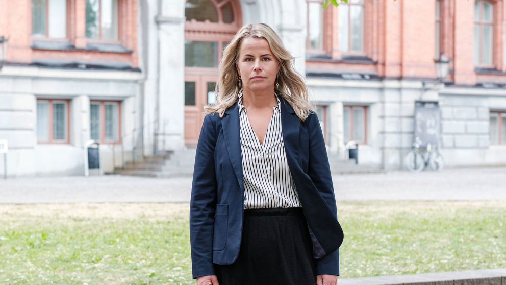 Sveriges Lärares ordförande Anna Olskog står utomhus på sommarhalvåret. Hon har neutral, nästan sur min och är klädd i vit blus med mörka ränder och en mörk kavaj.