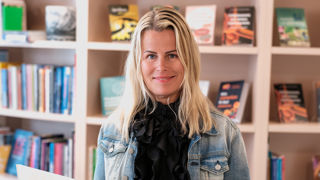 Sveriges Lärares ordförande Anna Olskog i svart tröja och blå jeansjacka ler och läser en bok med en bokhylla i bakgrunden.