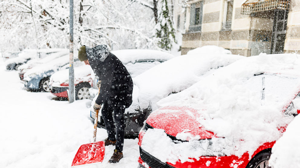 A Man with Snow Shovel is Shoveling a Snow in Front of Car During a Cold Snowy Winter Day.