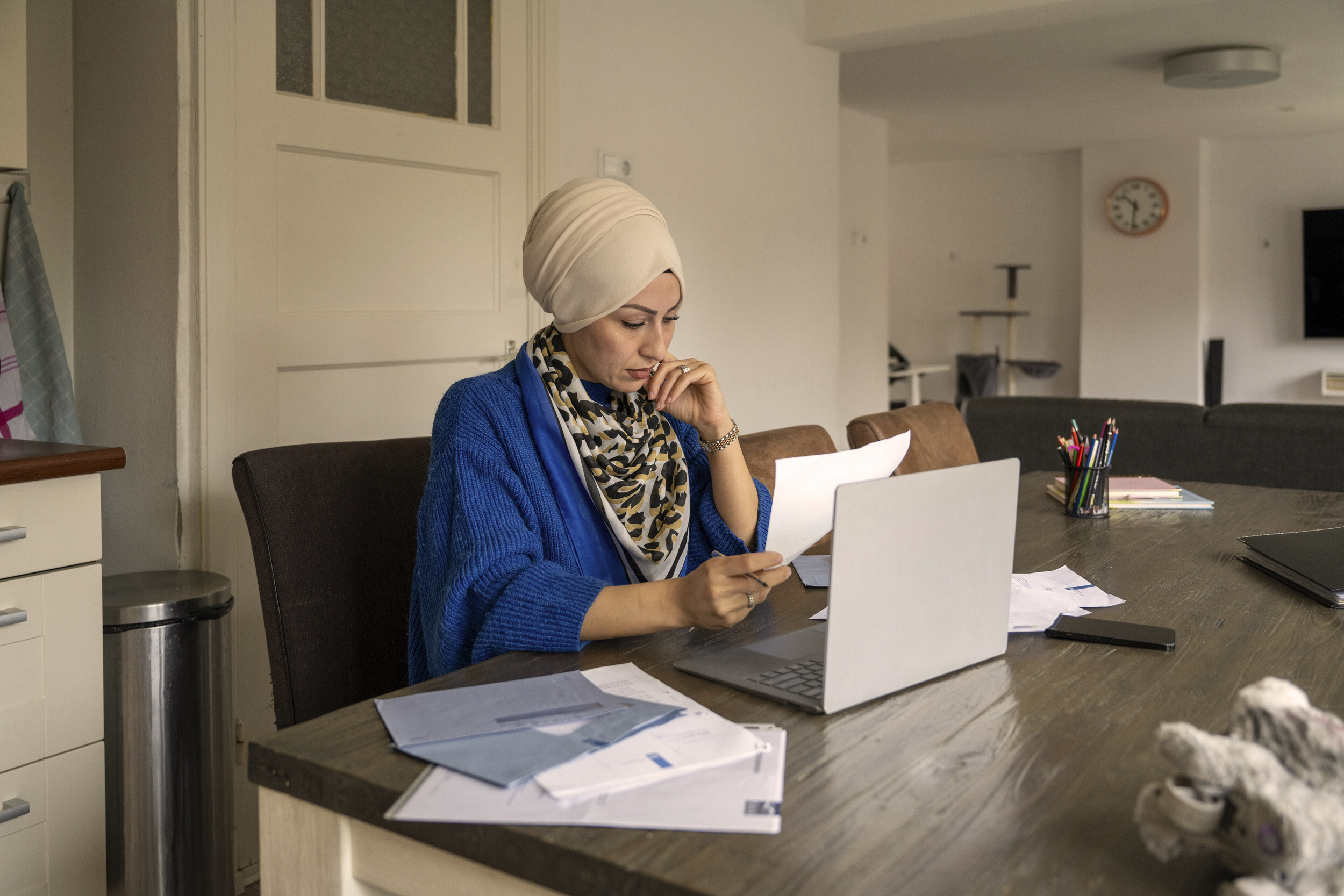 Adult woman wearing a hijab working on a laptop at a home office, thoughtfully holding a pen and analyzing documents. Adult woman wearing a hijab working on a laptop at a home office, thoughtfully holding a pen and analyzing documents. Adult woman wearing a hijab working on a laptop at a home office, thoughtfully holding a pen and analyzing documents.