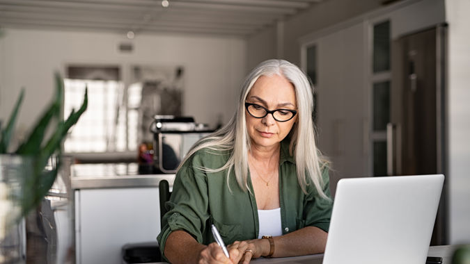 Senior stylish woman taking notes in notebook while using laptop at home. Old freelancer writing details on book while working on laptop in living room. Focused cool lady writing notary in notepad.