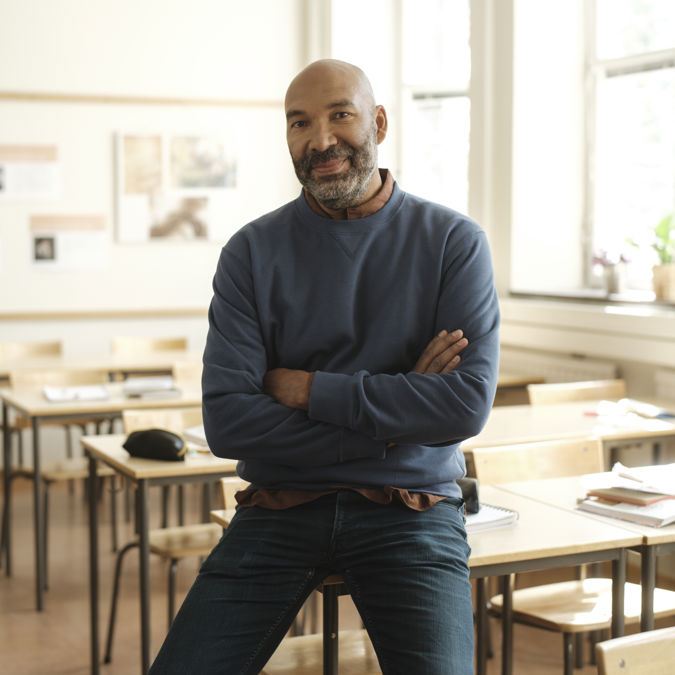 Leende manlig gymnasielärare halvsitter på en bänk i ett klassrum.
